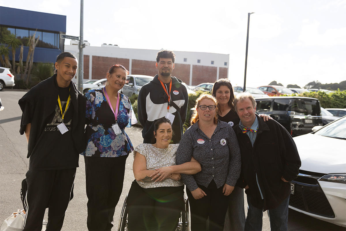 A group of seven people posing together outdoors in a parking lot, smiling at the camera, including one person seated in the front center.