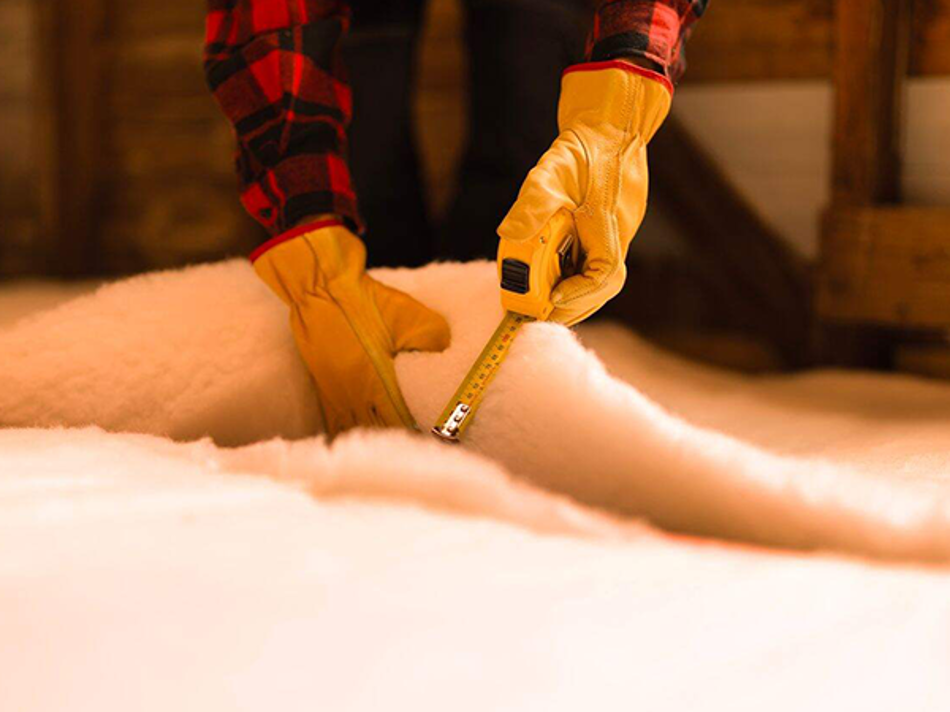 Hands in yellow work gloves using a tape measure on thick white insulation material.