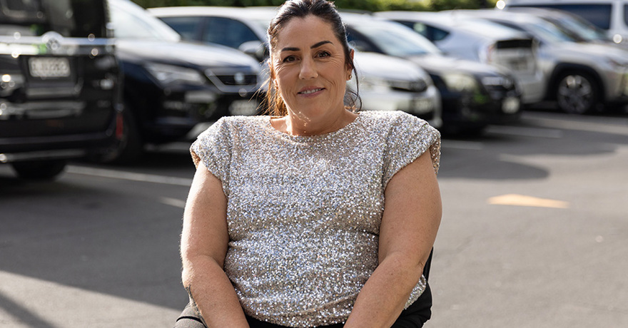 A smiling person wearing a glittering silver top and black trousers, seated outdoors in front of a parking lot filled with cars.