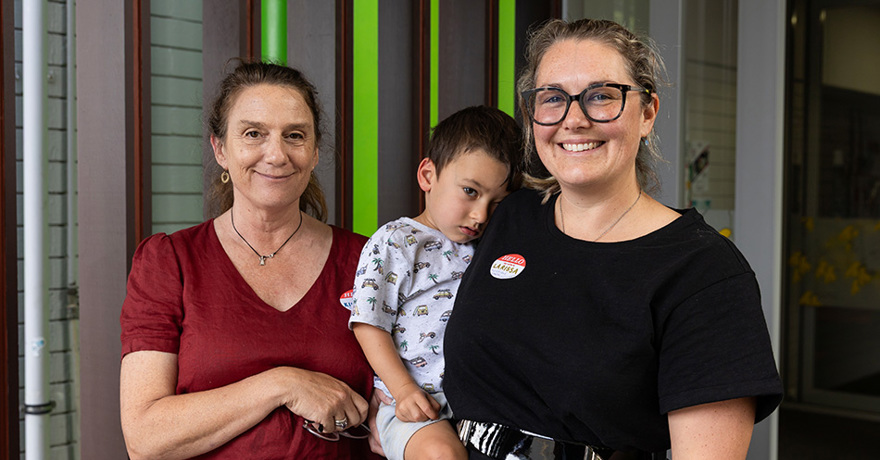 Two smiling adults standing together outdoors, with one holding a young child who is leaning against them. One adult wears a red shirt, and the other wears glasses, a black shirt, and a "Hello My Name Is Larissa" name tag.