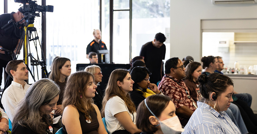 A diverse audience sits in rows listening attentively during a presentation or event. A large video camera on a tripod is visible in the upper left background near a window.