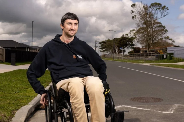 A smiling man in a black hoodie and beige trousers sitting in a wheelchair on a suburban street curb under a cloudy sky.