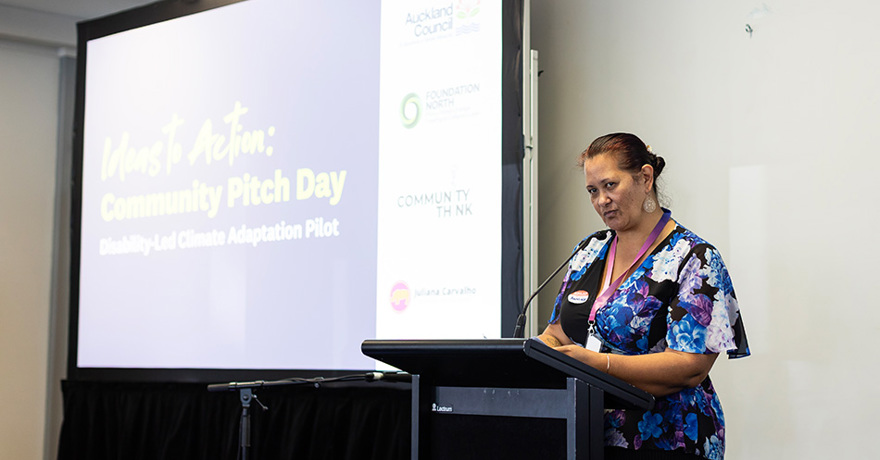 A person in a blue and purple floral top stands at a podium giving a presentation. Behind them, a large screen displays the text "Ideas to Action: Community Pitch Day – Disability-Led Climate Adaptation Pilot" alongside organizational logos.