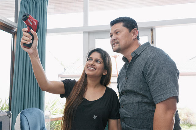 A smiling woman and a man using an infrared thermal imaging camera inside a home to check for drafts.