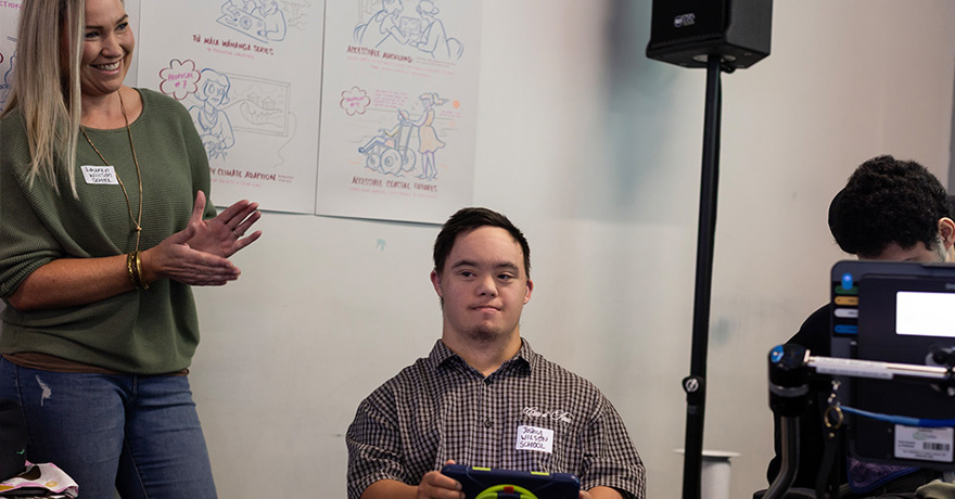 A person in a checkered shirt sits holding a tablet, while another person stands nearby smiling and clapping. In the background, illustrated posters about accessibility and climate adaptation hang on the wall next to a large speaker stand.