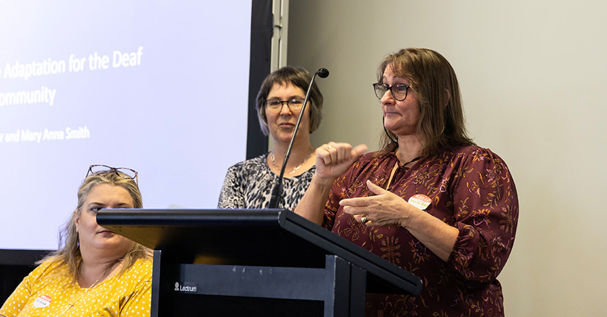 A person stands at a podium using sign language during a presentation, while another person stands behind them next to a projection screen and a third person sits listening on the left.