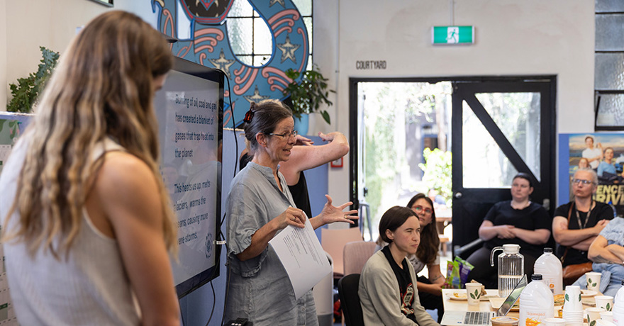 A person speaks and gestures with papers in hand during a presentation about climate change in a community space, while an audience listens around a table with refreshments.