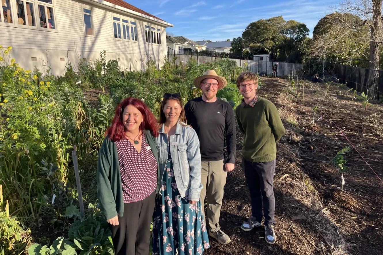 Four people stand smiling in a sunny garden, with leafy vegetables and yellow flowers on one side and freshly mulched beds with young plants on the other, in front of a white weatherboard building.