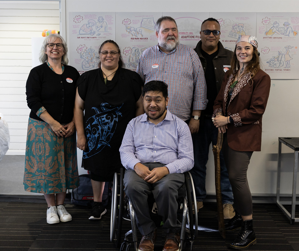 A group portrait of six people posing indoors. In the center foreground, a man in a striped shirt sits in a wheelchair, smiling.