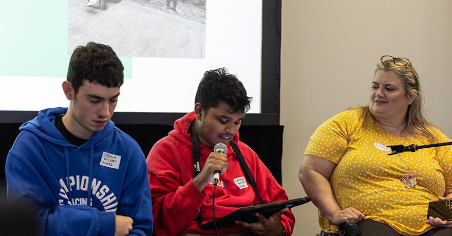 Three people are seated at a panel or presentation in front of a projection screen. The person in the center speaks into a microphone while looking at a tablet, flanked by a person in a blue hoodie on the left and a person in a yellow patterned shirt on t