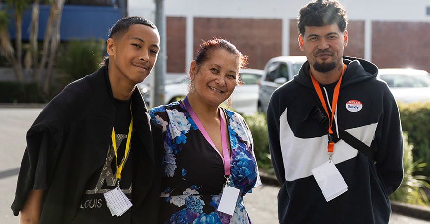 Three people stand together smiling outdoors in front of a building and parking area. They are all wearing lanyards with event badges, and the person on the right wears a black hoodie with a "Hello My Name Is Rocky" name tag.
