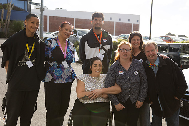 A group portrait of seven people standing and sitting closely together outdoors in a sunlit parking lot. In the front center, one individual in a textured white top sits in a wheelchair, with another individual standing beside them with an arm linked. Five other colleagues stand around them, all smiling at the camera. They wear casual attire, and several have lanyards or name tags. Cars and buildings are visible in the background.