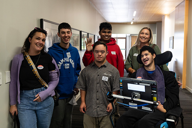 A group of six smiling individuals stands in a well-lit hallway. In the foreground, one person sits in a wheelchair with a communication device attached to the front, while another individual in a checkered shirt stands next to them. Behind them, two people stand on the left—one wearing a purple cardigan and the other in a blue hoodie—and two people stand on the right—one waving in a red hoodie and another smiling in a green sweater. Framed artwork hangs along the wall in the background.