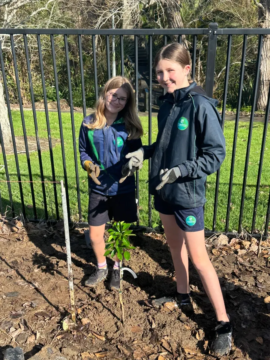 Two smiling students in school uniform and gardening gloves stand beside a small, newly planted leafy tree, in front of a black fence and lawn.