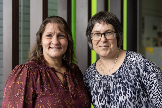 A close-up portrait of two women standing side-by-side indoors, smiling warmly at the camera. The woman on the left has light brown hair and is wearing a maroon floral blouse. The woman on the right has short dark hair, wears glasses, a silver chain necklace, and a black-and-white patterned top. The background features vertical wooden slats and a bright green accent wall.