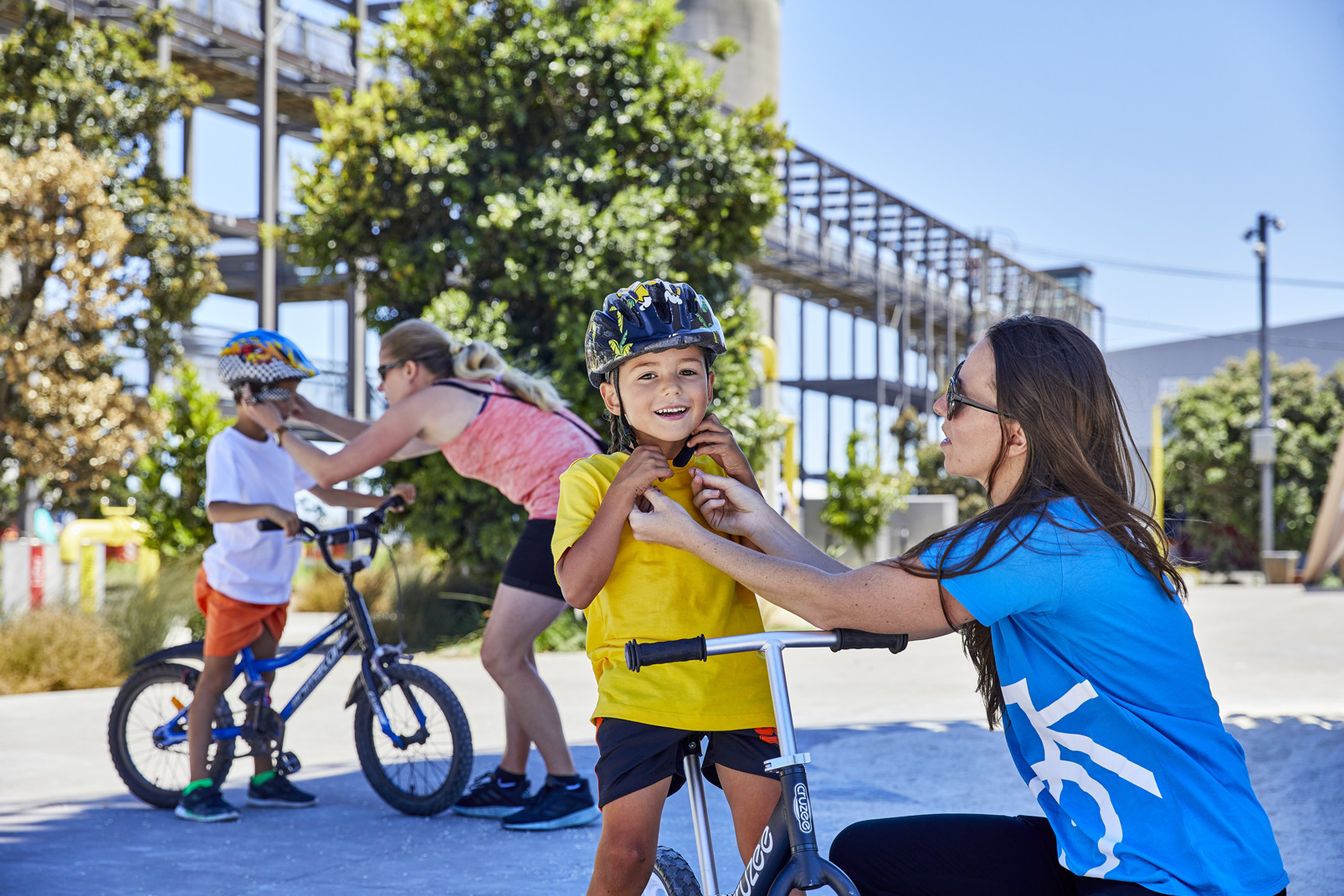 A coach helps a smiling child fasten their bike helmet at a learn-to-ride session.